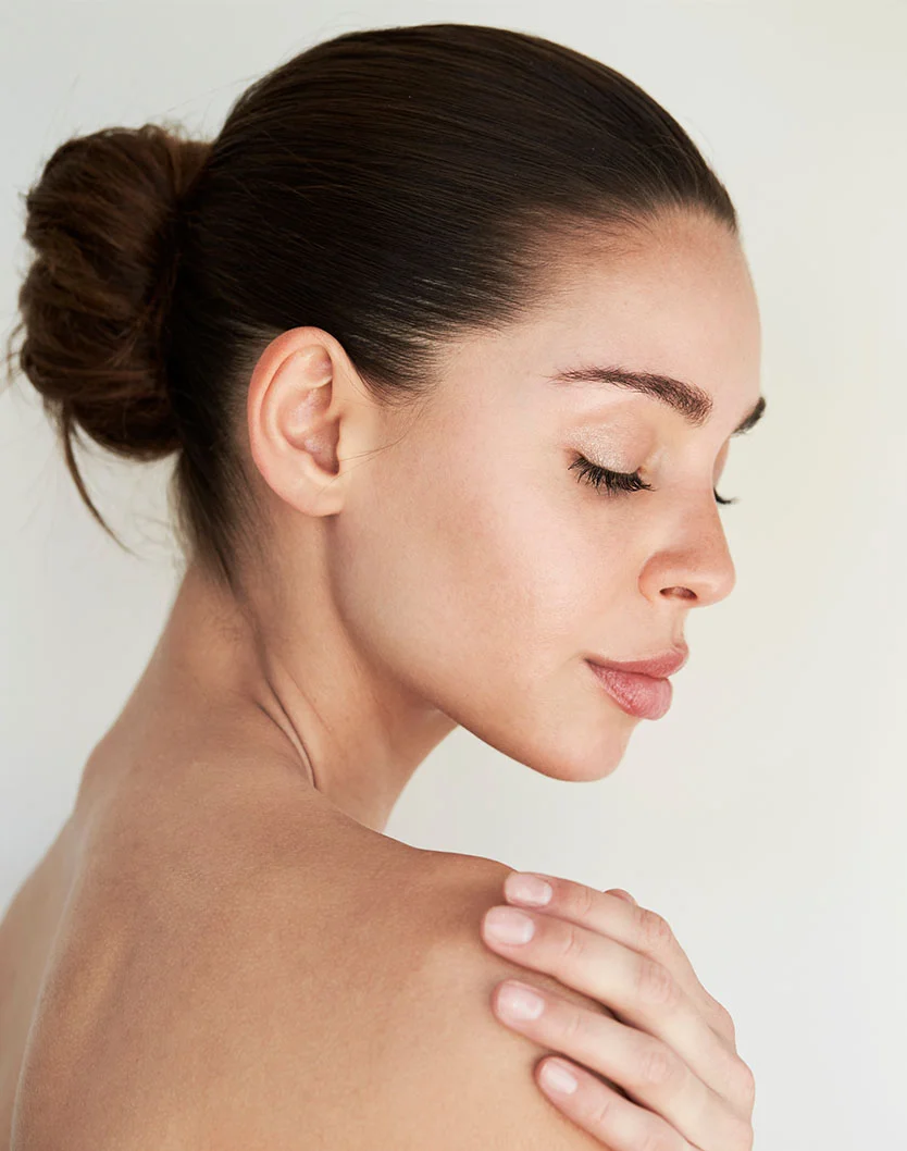Close-up of a woman with light-toned skin and dark brown hair in a loose bun. Her eyes are closed, and she faces slightly right with a calm expression. Her hands rest gently on her shoulder. The background is a plain, light beige, emphasizing her smooth skin and serene demeanor. - Back Lift in Fairfax, VA