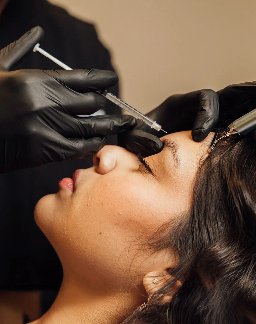 Close-up of a person receiving a cosmetic injection. They are lying down with their face angled toward the camera, eyes closed, and mouth slightly open. A professional in black medical gloves is administering the injection to their forehead. The focus is on the face and gloved hands, with background elements like clothing and a medical device softly blurred. - Botox for Wrinkle Relaxers in Fairfax, VA