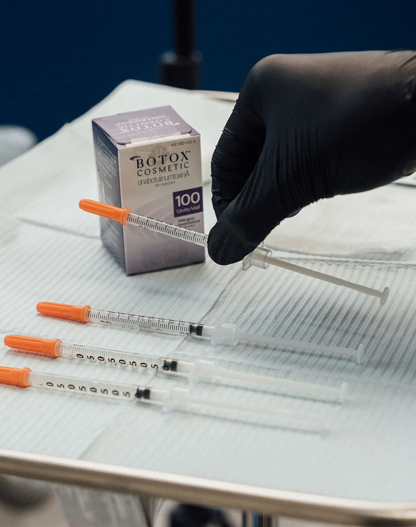 Close-up of a stainless steel tray holding a box of Botox Cosmetic and several syringes with orange caps. A gloved hand in black nitrile gloves holds one syringe. The background is a muted blue-gray and out of focus. - Botox for Calf Reduction in Fairfax, VA