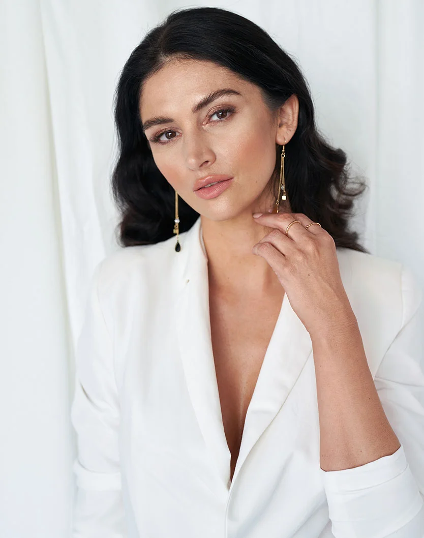 A light-skinned woman with dark shoulder-length hair and subtle makeup wears a white collared blazer-style top with a V-neckline. She rests her hand gently on her throat, showing a thin ring. Long gold earrings with dark stones hang from her ears. The background is plain off-white, with soft, even lighting highlighting her skin and clothing. - Browlift in Fairfax, VA