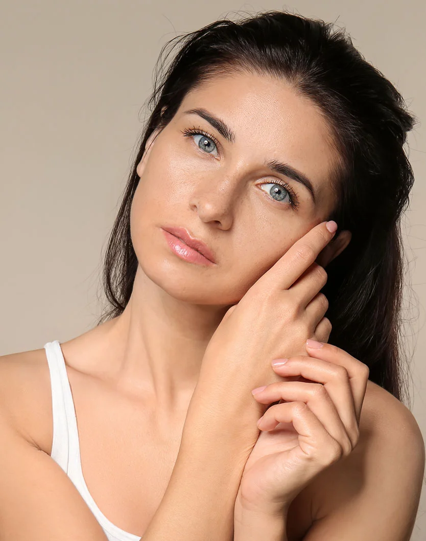 A close-up of a woman with long dark hair and light, smooth skin. She wears a white tank top and has a calm, neutral expression, looking slightly to the right. Her hands gently touch her face and neck, with light pink nails. The muted beige background softly highlights her features. - Dermaplaning in Fairfax, VA