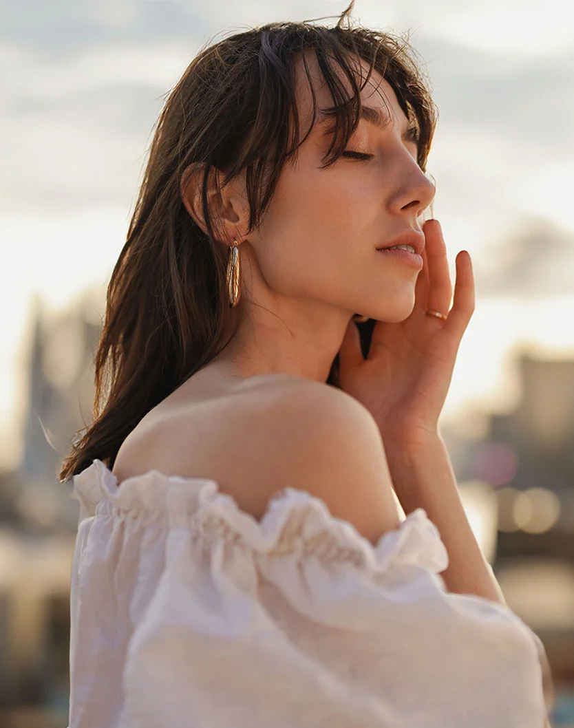 Close-up portrait of a woman with shoulder-length dark brown hair, eyes closed and a serene expression, gently touching her cheek. She wears a white off-the-shoulder top with ruffled detailing and gold hoop earrings. Warm, golden-hour lighting highlights her face, with a softly blurred urban background. - Facial Fat Grafting in Fairfax, VA