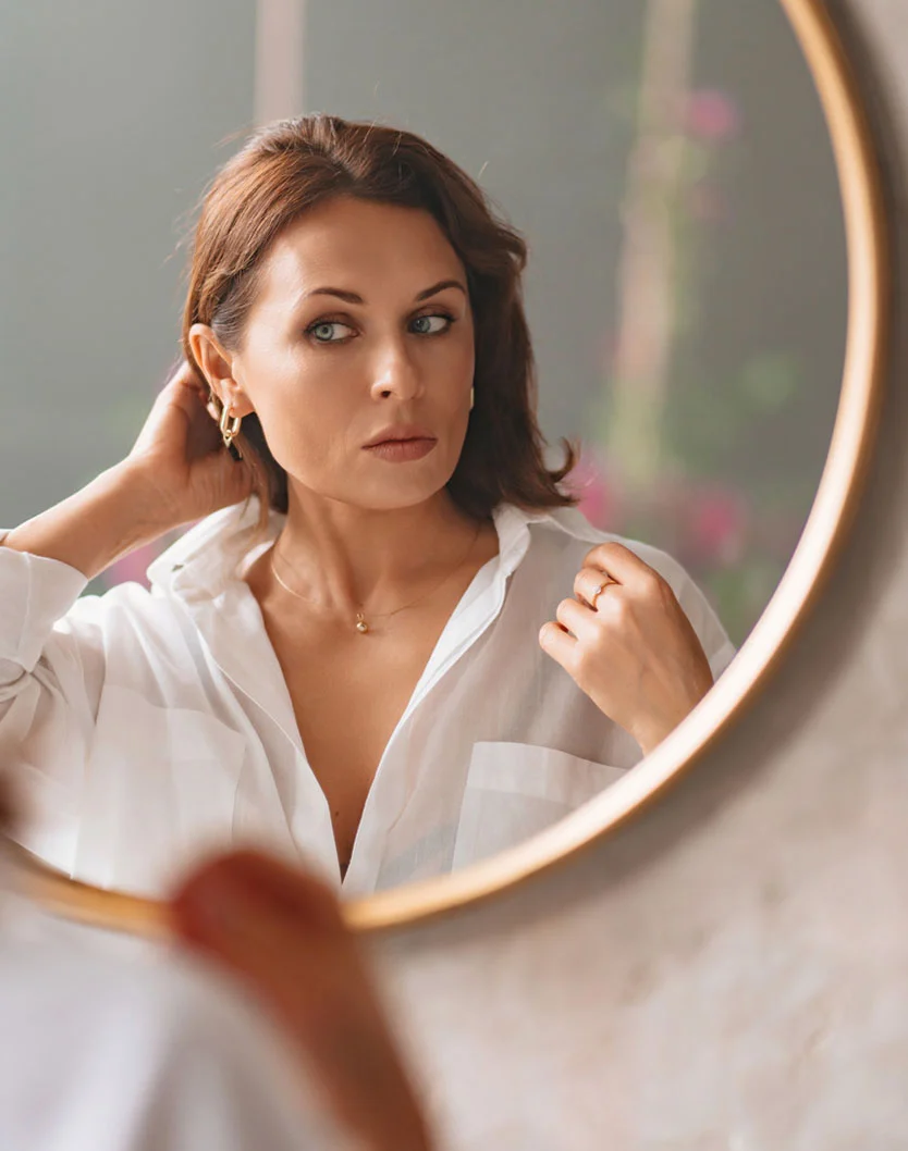 A woman with shoulder-length brown hair adjusts her hair and white button-down shirt while looking into a round, gold-framed mirror. She has a focused expression and wears a delicate gold necklace and ring. The mirror reflects a soft, light-gray or beige room, with part of another person's arm visible. - Hair Transplant in Fairfax, VA