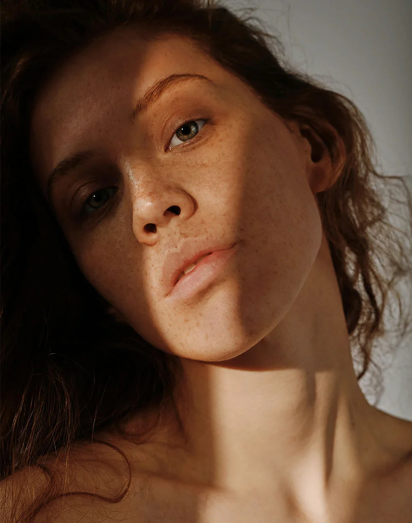 Close-up of a woman with shoulder-length, wavy dark brown hair and light tan skin with freckles across her nose and cheeks. Soft lighting casts shadows that highlight her features. She has light green eyes and natural rosy pink lips. The background is softly blurred, emphasizing her face and neck in a natural, gentle style. - Medical Grade Facials in Fairfax, VA