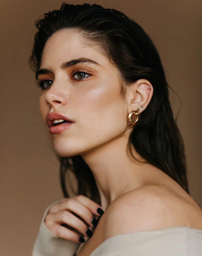 Close-up portrait of a woman with dark, slightly damp hair framing her shoulders. She has smooth, light-toned skin with subtle blush and highlight on her cheekbones. Her hazel-green eyes look ahead, and her natural pink lips are slightly parted. She wears gold hoop earrings, a delicate chain necklace, and a light beige off-the-shoulder sweater. Her nails are painted black. The background is a muted light brownish-tan, creating a soft, natural beauty aesthetic. - Microneedling in Fairfax, VA
