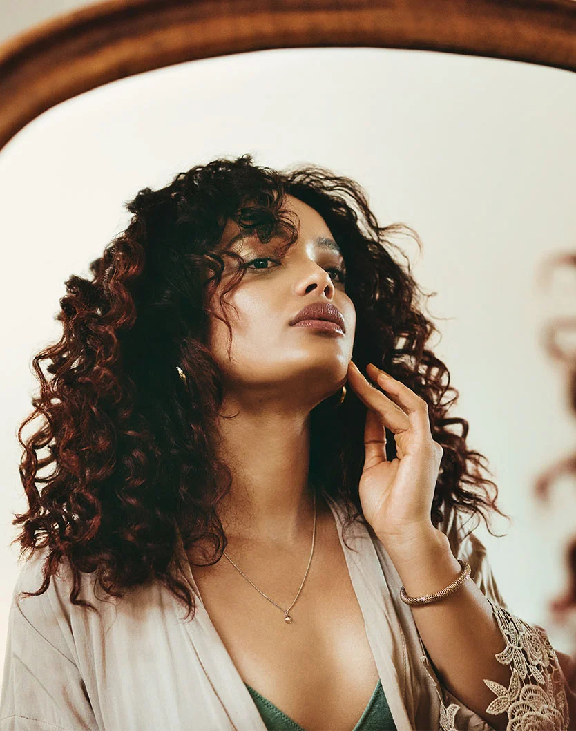 A woman with dark, curly hair gazes thoughtfully at her reflection in a mirror. She wears a loose beige robe over a muted teal-green top, a delicate silver necklace with a small pendant, and a bracelet. Her makeup is subtle with shimmery gold eyeshadow and nude lipstick. She gently touches her cheek, framed by a warm light brown mirror frame in a softly lit indoor setting. - Necklift in Fairfax, VA