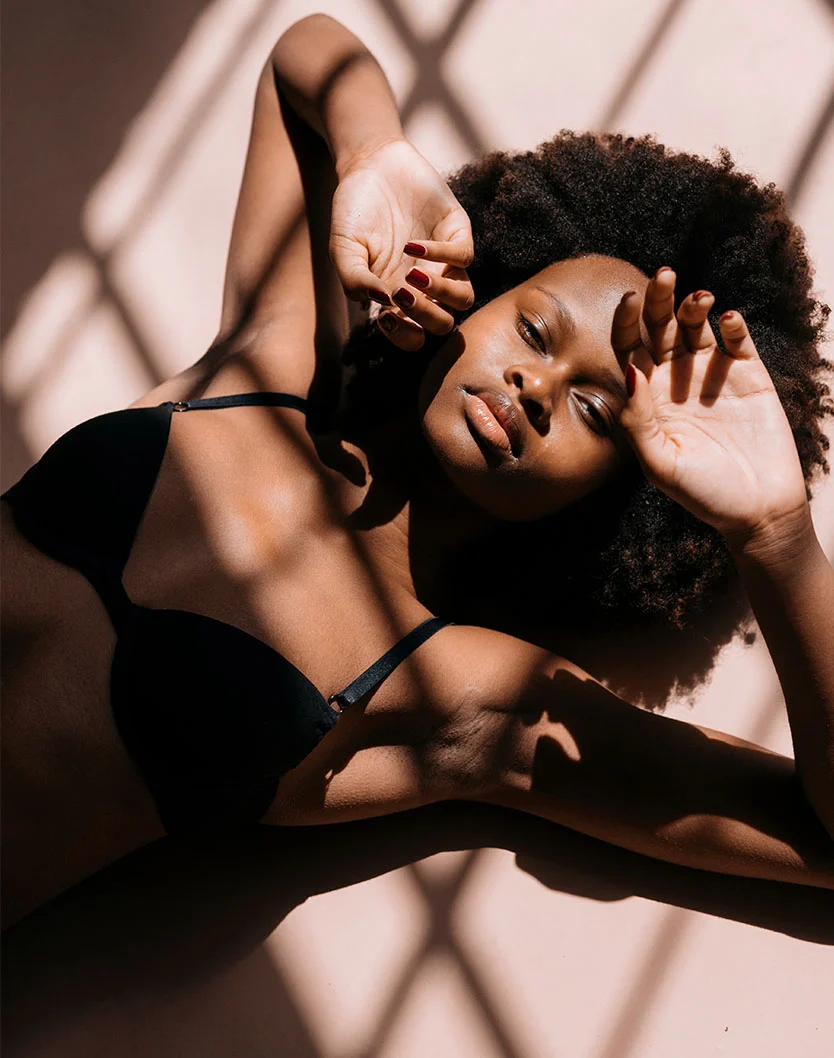 High-angle, full shot of a dark-skinned woman lying on a light peach surface, wearing a black bra. Her arms are outstretched with hands resting on her face, partially covering her eyes. Her voluminous, naturally curly dark brown hair fans around her head. Crisscrossing shadows from a window fall across her body and the background. Her expression is calm and restful. - Nipple Reduction in Fairfax, VA