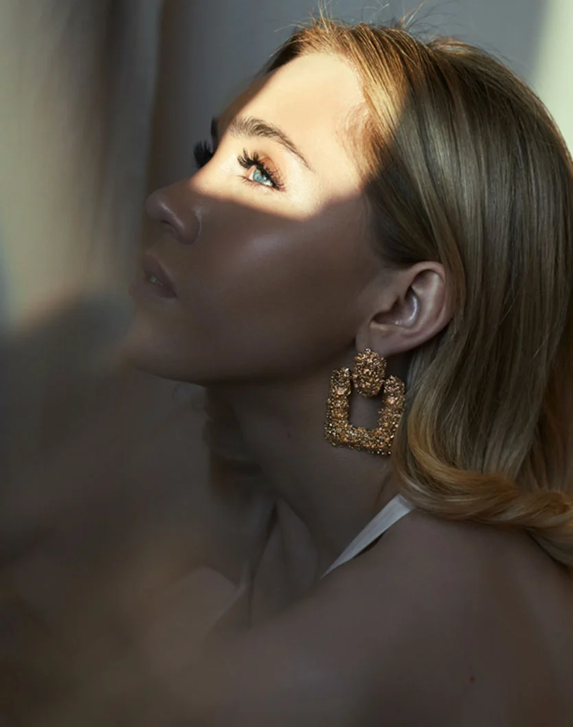 Close-up profile of a woman with light blonde, wavy hair. Her smooth, tanned skin is highlighted by sunlight casting shadows across her face, emphasizing her light blue eye and cheekbones. She wears large, textured gold earrings with clustered detailing. A soft, white fabric is visible around her neck and shoulders. - Permanent Makeup in Fairfax, VA