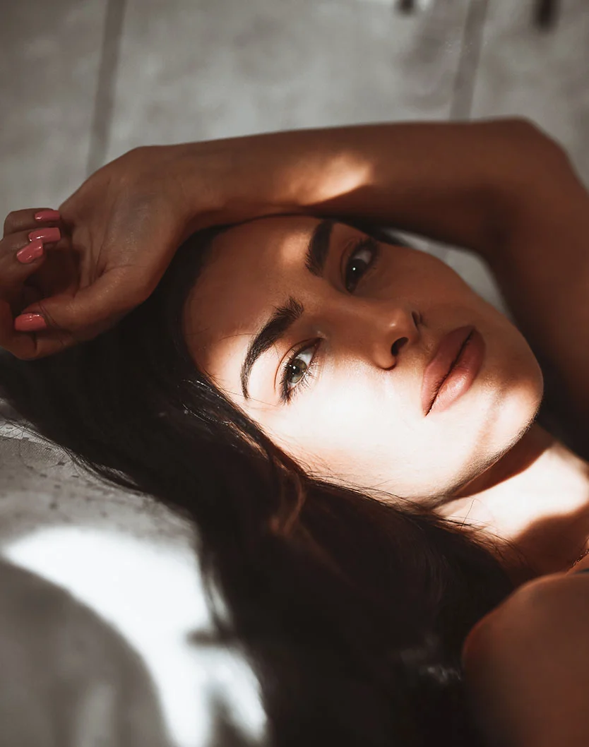 A close up shot of a womans face glancing towards the camera with one arm rested above her head. She is laying down with a textured wall seen in the back. - Rhinoplasty in Fairfax, VA