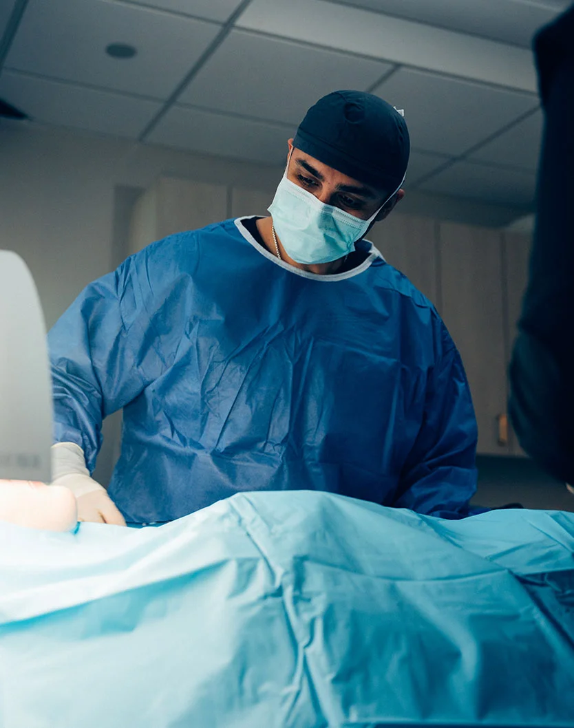 Dr. Chopra preforming surgery, wearing scrubs, a mask and gloves. Showing his enthuses for a clean and calm procedure. - Tummy Tuck in Fairfax, VA